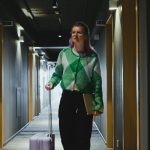 Woman walking through hotel corridor with suitcase, wearing green cardigan.