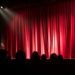 Dimly lit theater stage with red curtains and audience silhouettes under spotlights.