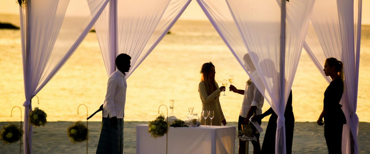 Elegant beach wedding ceremony setup with guests during a vibrant sunset on the waterfront.